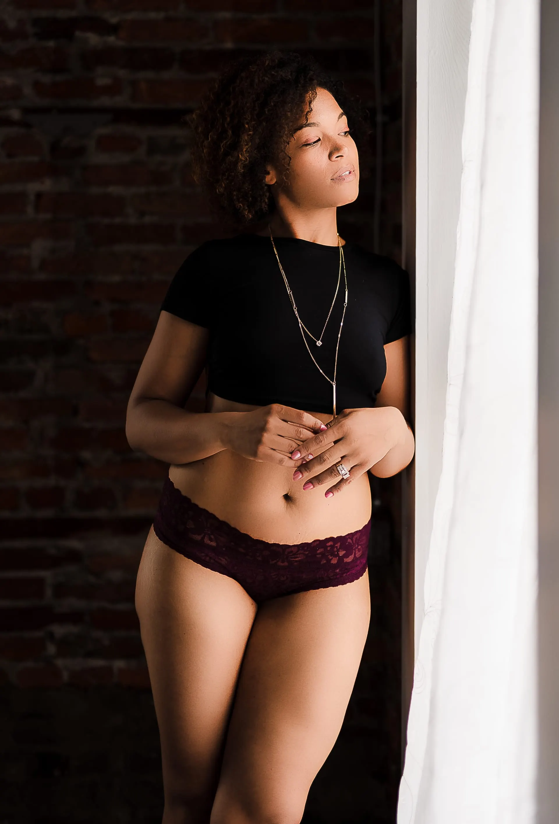 Woman in a black crop tee and burgundy lace briefs leaning by a window
