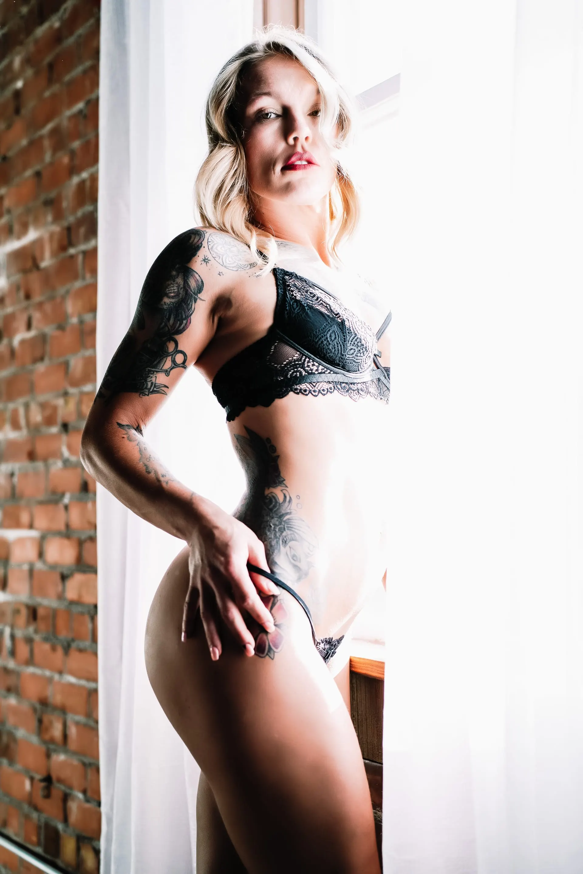 Backlit portrait in black lace beside a brick wall and white curtains