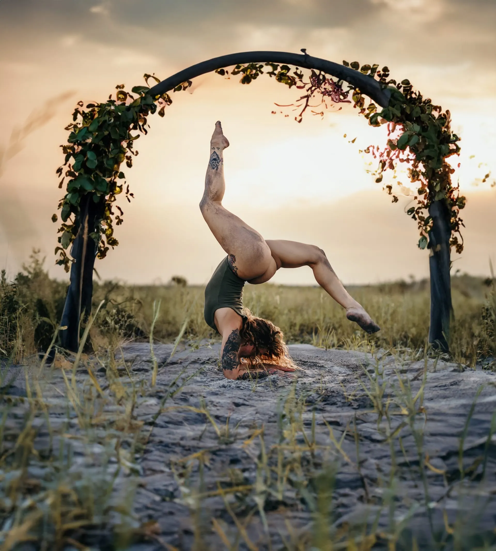 Handstand in an olive bodysuit beneath a vine-covered arch in an open field